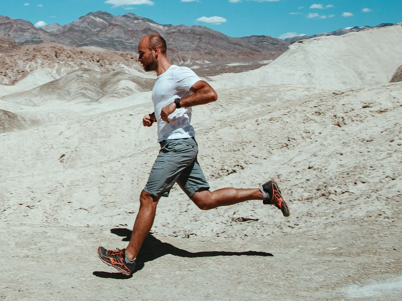 A runner in a white t-shirt running along a desert trail with mountains behind him